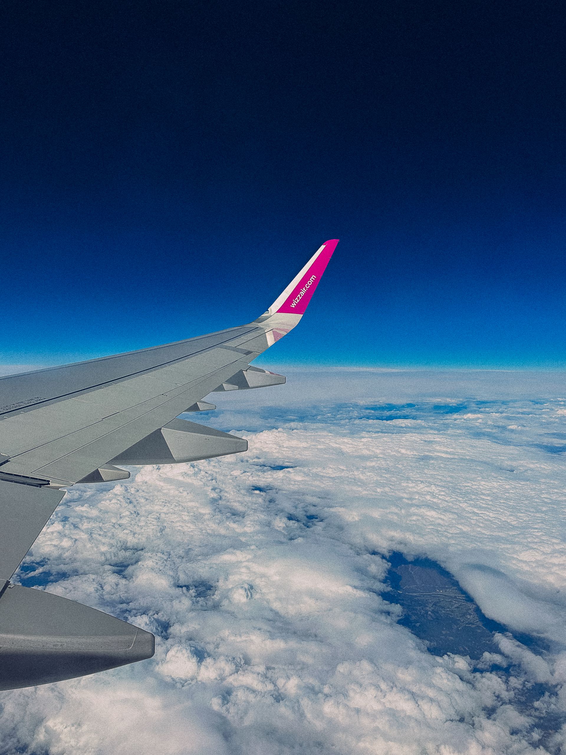 View of an airplane wing flying over clouds with clear blue sky.